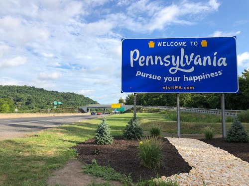 Welcome sign for Pennsylvania with the slogan "pursue your happiness," surrounded by greenery and a highway.