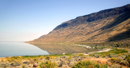 A serene landscape featuring a calm lake reflecting a rugged mountain and a sandy shore, surrounded by sparse vegetation.