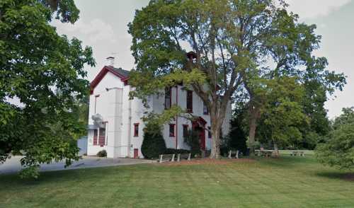 A white two-story building surrounded by trees and a grassy area, with benches and a clear sky in the background.