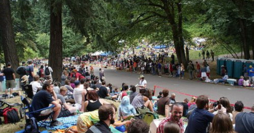 A large crowd gathers along a road in a forested area, with people sitting on the ground and portable restrooms nearby.