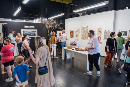 A lively art gallery event with people mingling, viewing artwork, and enjoying refreshments in a bright, spacious setting.