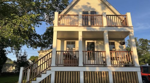 A two-story house with a porch, wooden railings, and stairs, surrounded by trees and a clear blue sky.