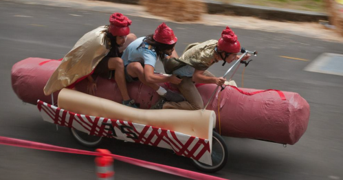 Three people in costumes riding a bicycle designed to look like a giant hot dog, racing down a street.