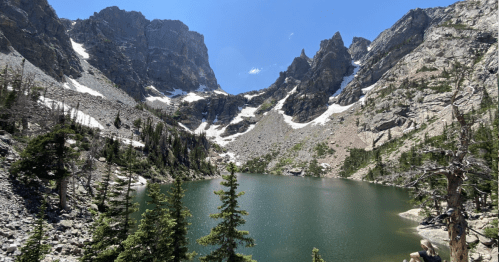 A serene mountain lake surrounded by rocky peaks and pine trees under a clear blue sky. Snow patches visible on the slopes.