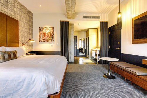 A modern hotel room featuring a large bed, artwork, and a stylish seating area with a view of the bathroom.