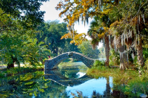 A stone bridge arches over a calm pond, surrounded by lush greenery and hanging moss in a serene park setting.