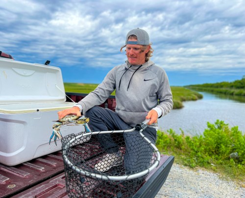 A person in a gray hoodie holds a crab over a net by a river, with a cooler and greenery in the background.