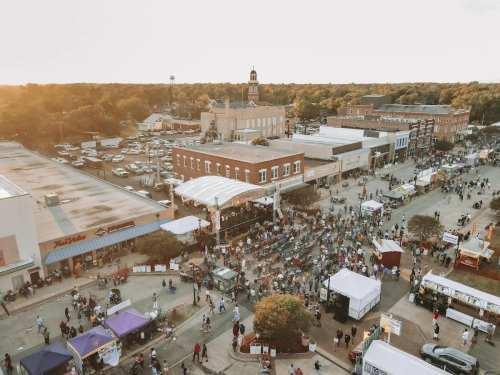 Aerial view of a bustling street fair with vendors, crowds, and historic buildings under a warm sunset.