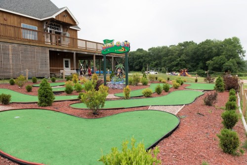 A mini-golf course with green turf and landscaped areas, located near a wooden building and playground.