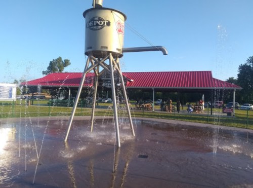 A large water fountain shaped like a vintage water tower, with water spraying around it in a park setting.