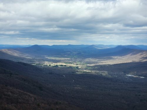 A panoramic view of a valley surrounded by mountains under a cloudy sky, with patches of green and brown landscape.