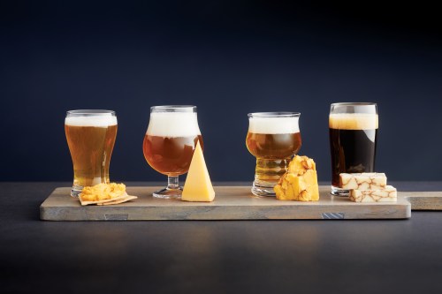 A tasting board featuring four glasses of beer and various cheeses on a slate platter against a dark background.