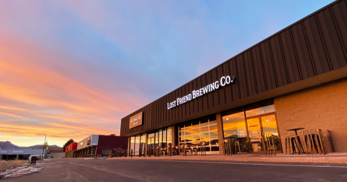 Exterior of Lost Friend Brewing Co. at sunset, showcasing a modern building with large windows and a colorful sky.