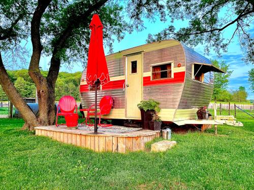A vintage silver trailer with red accents sits on a wooden deck under a tree, surrounded by green grass and red chairs.