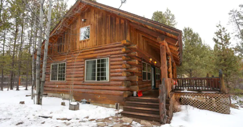 A rustic log cabin surrounded by snow and trees, featuring a porch and large windows.
