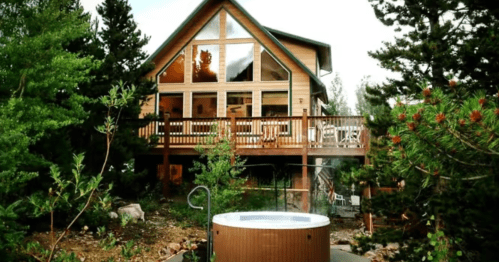 A cozy cabin with large windows, a deck, and a hot tub surrounded by trees and greenery.