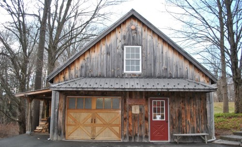 A rustic wooden cabin with a metal roof, featuring a garage door and a red front door, surrounded by bare trees.