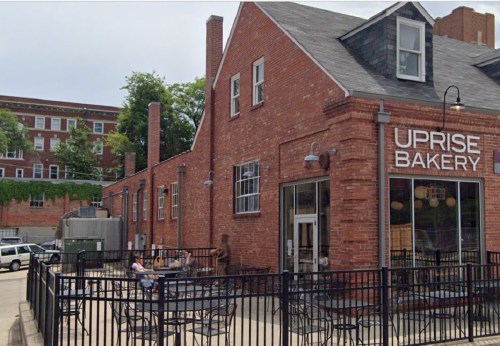 Exterior of Uprise Bakery featuring a brick building, outdoor seating, and nearby greenery.
