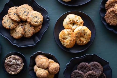 A variety of cookies and baked goods arranged on black plates, including chocolate chip, snickerdoodles, and bagels.
