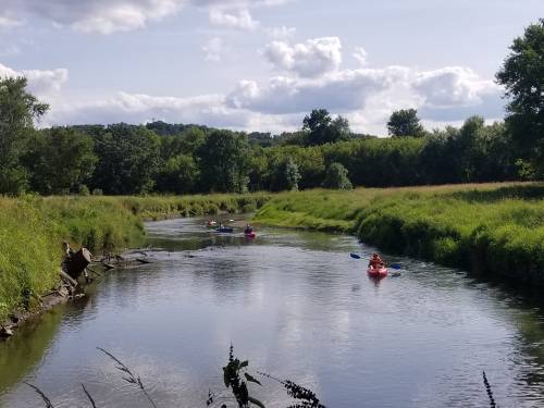 A serene river scene with kayakers paddling through lush greenery under a partly cloudy sky.
