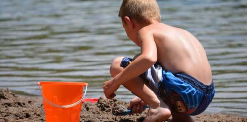 A young boy crouches by the water, playing in the sand with a small orange bucket.