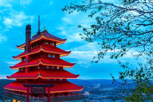 A vibrant red pagoda stands against a blue sky, illuminated at dusk, surrounded by trees and a scenic view.