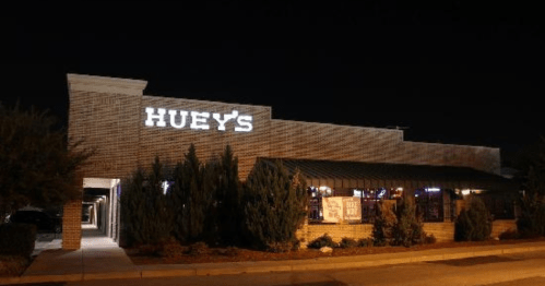 A brick restaurant exterior at night with the sign "HUEY'S" illuminated, surrounded by greenery.