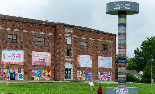 A colorful mural on a brick building featuring the "World's Largest Spool of Thread" sign and various quilt designs.