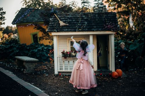 A child in a pink dress and fairy wings stands by a decorated playhouse, holding a wand, with pumpkins nearby.
