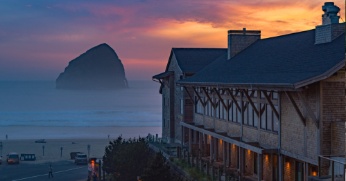 A coastal scene at sunset featuring a beach, a large rock formation, and a wooden building with warm lights.