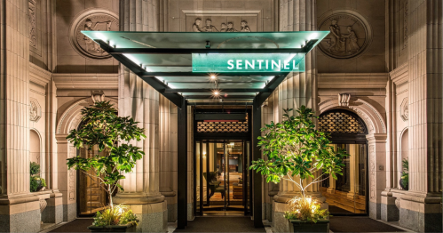 Entrance of the Sentinel building, featuring a glass canopy and decorative plants, illuminated at night.
