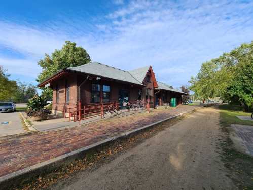 A brick train station with a sloped roof, surrounded by trees and a pathway, featuring bicycles parked outside.