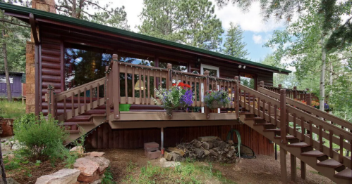 A cozy log cabin with a wooden deck, surrounded by trees and colorful flower pots, set in a natural landscape.