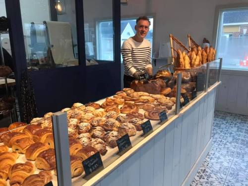 A baker stands behind a display of various pastries and breads in a cozy bakery setting.