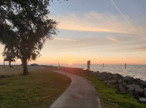A serene waterfront path at sunset, with trees and rocks lining the shore under a colorful sky.