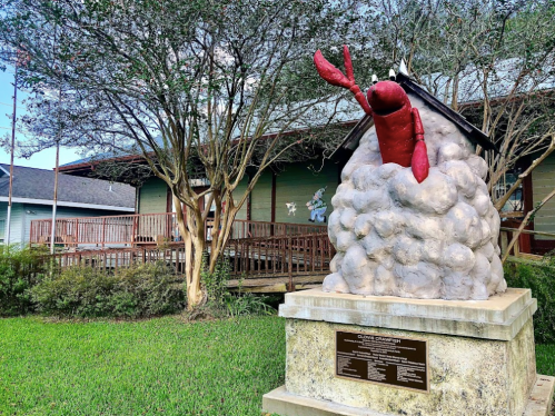 A whimsical lobster sculpture emerging from a cloud, set in a grassy area near a wooden building.