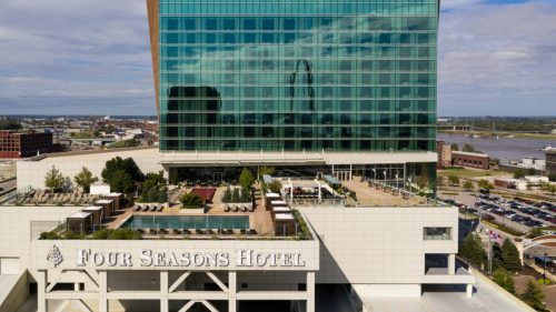 Aerial view of the Four Seasons Hotel with a modern glass facade and rooftop pool area, overlooking a cityscape.