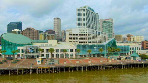 City skyline with modern buildings and palm trees along a waterfront promenade. Cloudy sky above.