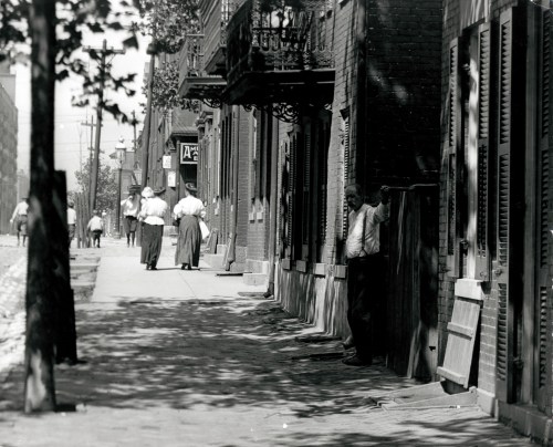 Black and white photo of a street scene with people walking, buildings, and shadows from trees.