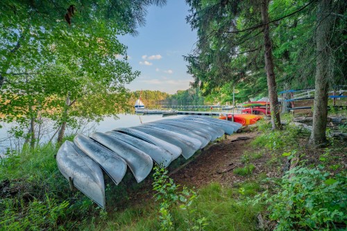 A row of canoes rests on the shore beside a calm lake, surrounded by trees and a distant dock.