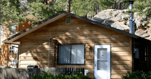 A cozy wooden cabin surrounded by trees and rocks, featuring a front porch and a white door.