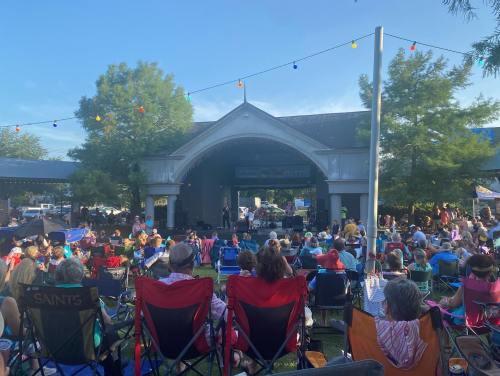 A crowd of people sitting in chairs at an outdoor concert, with a stage and colorful lights in the background.