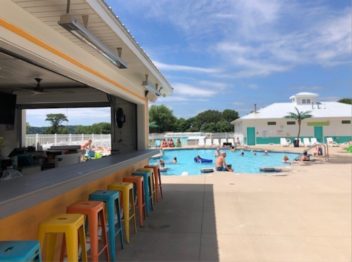 A sunny pool area with people swimming, colorful bar stools in the foreground, and a bar serving drinks.