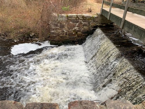 A small waterfall cascades over a stone wall, with frothy water flowing into a calm stream below.