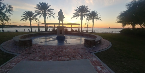 A statue stands in a circular plaza surrounded by palm trees, with a sunset over the water in the background.