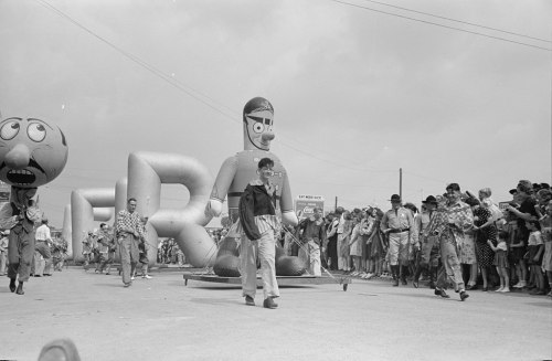 A black and white photo of a parade with large balloons and costumed characters, surrounded by a crowd of spectators.