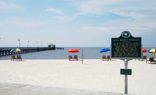 A beach scene with colorful umbrellas, lounge chairs, and a historical marker about civil rights wade-ins near a pier.