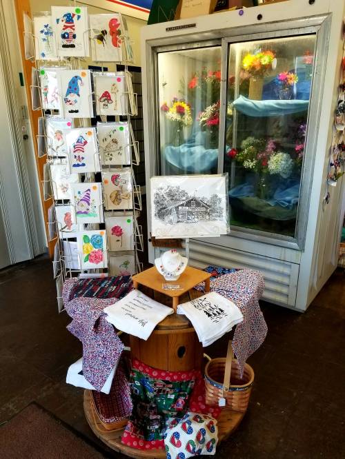 A display of handmade cards, art, and crafts in a shop, with a cooler of flowers in the background.