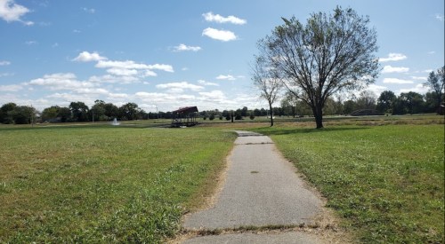 A paved path leads through a grassy park with a tree and a pavilion under a blue sky with scattered clouds.