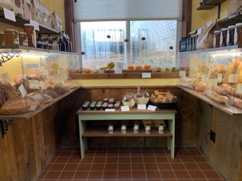 A cozy bakery display with various breads, pastries, and jars of preserves on wooden shelves and a table.